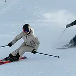 A skier in white winter gear and a black helmet skiing down fresh snow with a companion in the background, enjoying a winter day.