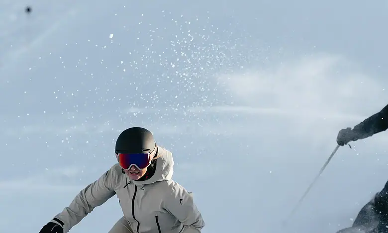 A skier in white winter gear and a black helmet skiing down fresh snow with a companion in the background, enjoying a winter day.