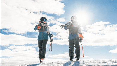 Two skiers in Halti outdoor clothing on snowy mountain, bright sun and blue sky in background.