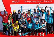 Group of winter sports athletes celebrating on the podium with medals and skis, in front of a red backdrop with Audi branding.