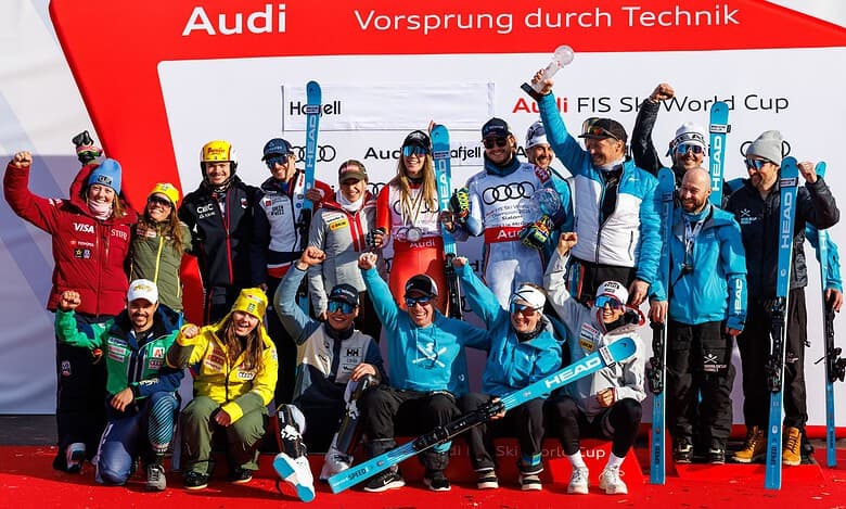 Group of winter sports athletes celebrating on the podium with medals and skis, in front of a red backdrop with Audi branding.
