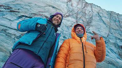 Two explorers in colorful winter jackets standing in front of a glacier, smiling and enjoying the winter outdoor adventure.