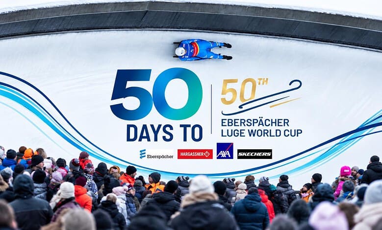 Crowd watching a luge race at the Eberspächer Luge World Cup with a large banner displaying '50 Days To' and event logos.