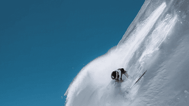 Skier descending a steep, snow-covered mountain slope during bright winter day, showcasing thrilling winter sports activity.