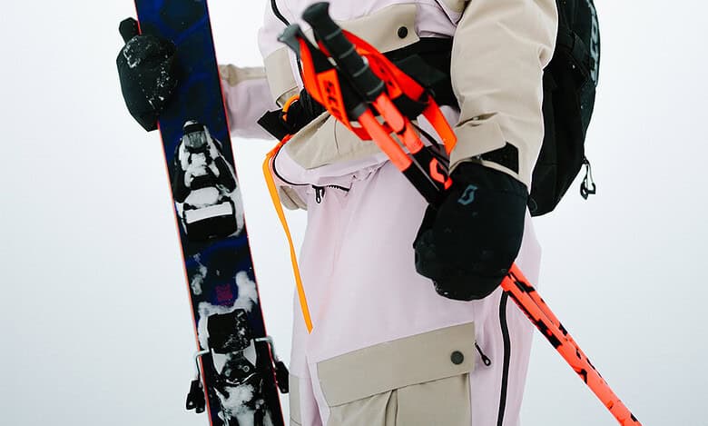 Snowboarder in winter gear holding a snowboard and ski poles, ready for winter sports on a snowy mountain.