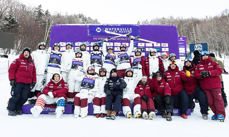 Group of winter sports athletes and coaches celebrating on snow with medals and trophies at Waterville Valley Resort.