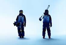 Two snowboarders in blue winter outfits standing on a pale blue background, one holding a snowboard and the other with a ski pole.