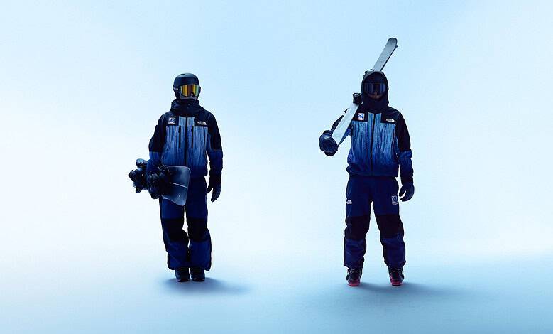 Two snowboarders in blue winter outfits standing on a pale blue background, one holding a snowboard and the other with a ski pole.