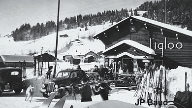 Vintage black and white winter scene at a ski resort with people, cars, and snow-covered buildings.