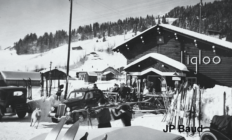Vintage black and white winter scene at a ski resort with people, cars, and snow-covered buildings.