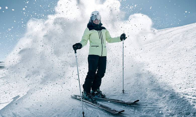 Skier in pale yellow jacket and black pants navigating fresh powder on a sunny snowy slope with blue sky.