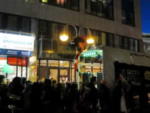 Bunter Demonstrationszug vor einem Geschäft mit Zelten und Flaggen bei Nacht.