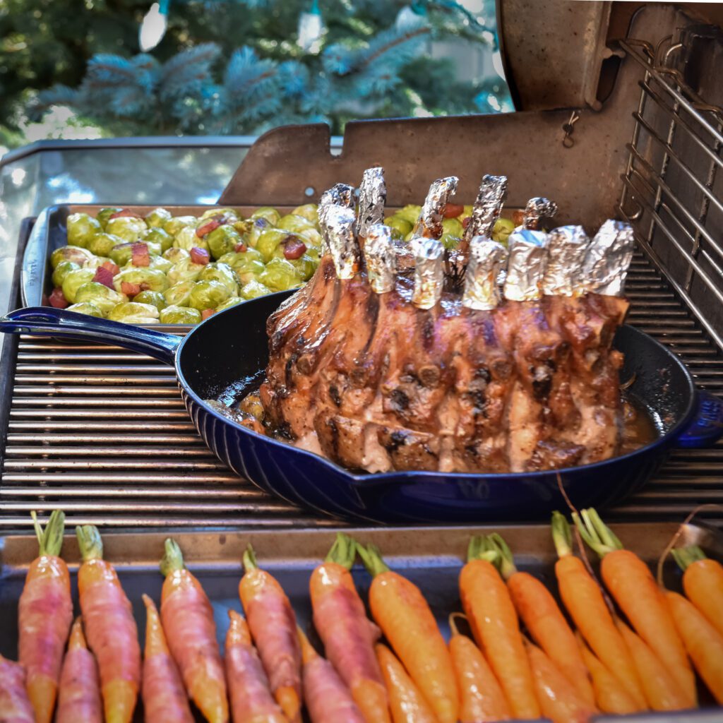 Raw crown roast of pork in a blue cast-iron pan with tin foil on the bones on the barbeque. Carrots and brussels sprouts also on the grill.