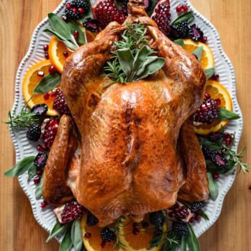 Dry-Brined & Grilled Turkey, top down view of a golden and grilled whole turkey on a platter and garnished with oranges, pomegranates and herbs.