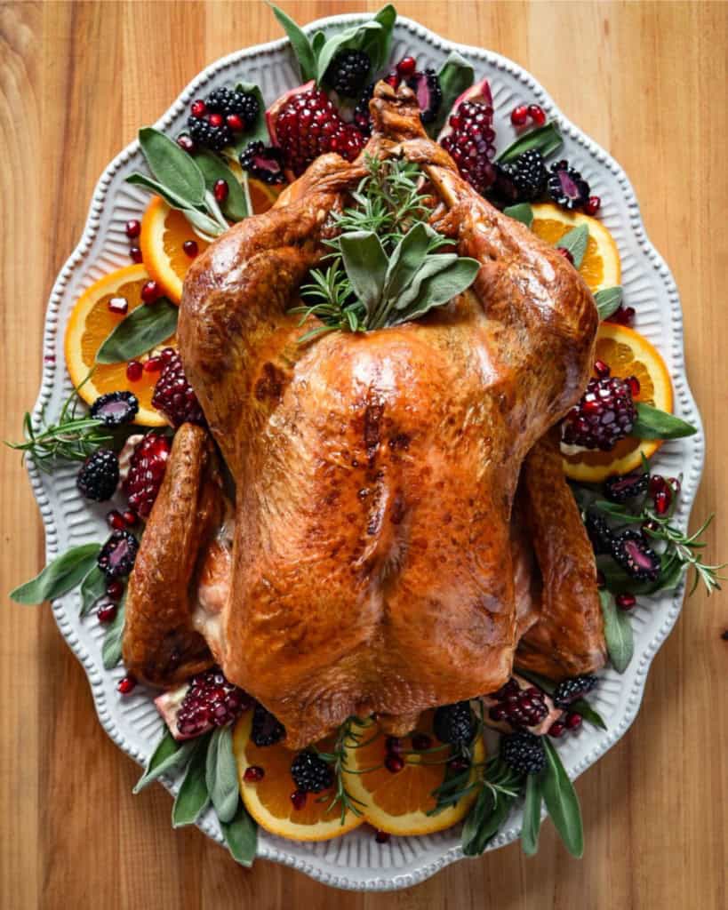 Dry-Brined & Grilled Turkey, top down view of a golden and grilled whole turkey on a platter and garnished with oranges, pomegranates and herbs.