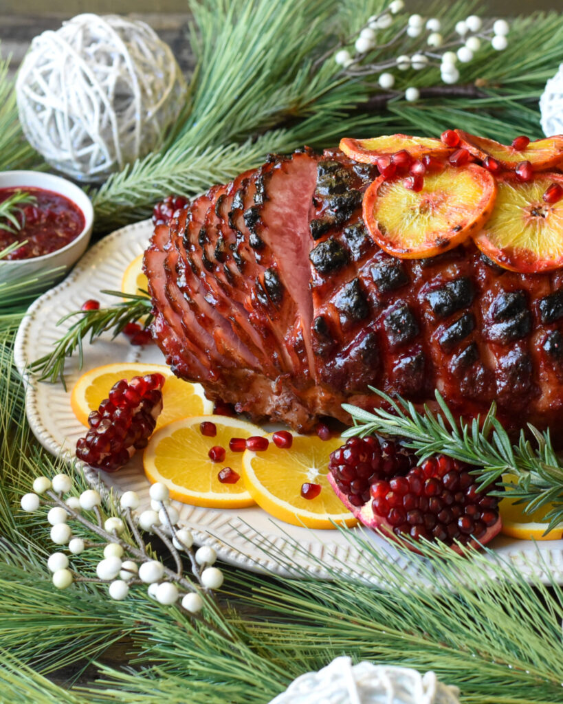 Side view shot of a glazed ham sliced garnished with pomegranates, rosemary and sliced oranges. Surrounded by Christmas decorations and a bowl of cranberry sauce.