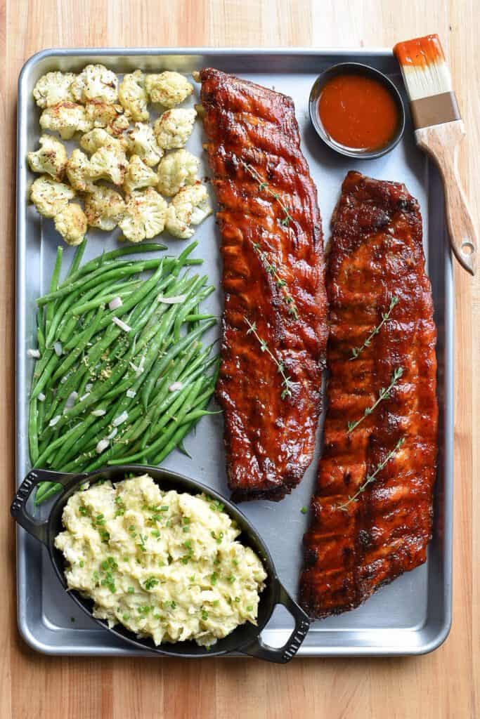 Sheet pan filled with two racks of ribs, cauliflour, green beans and mashed potatoes. Extra sauce in bowl with a brissel brush.