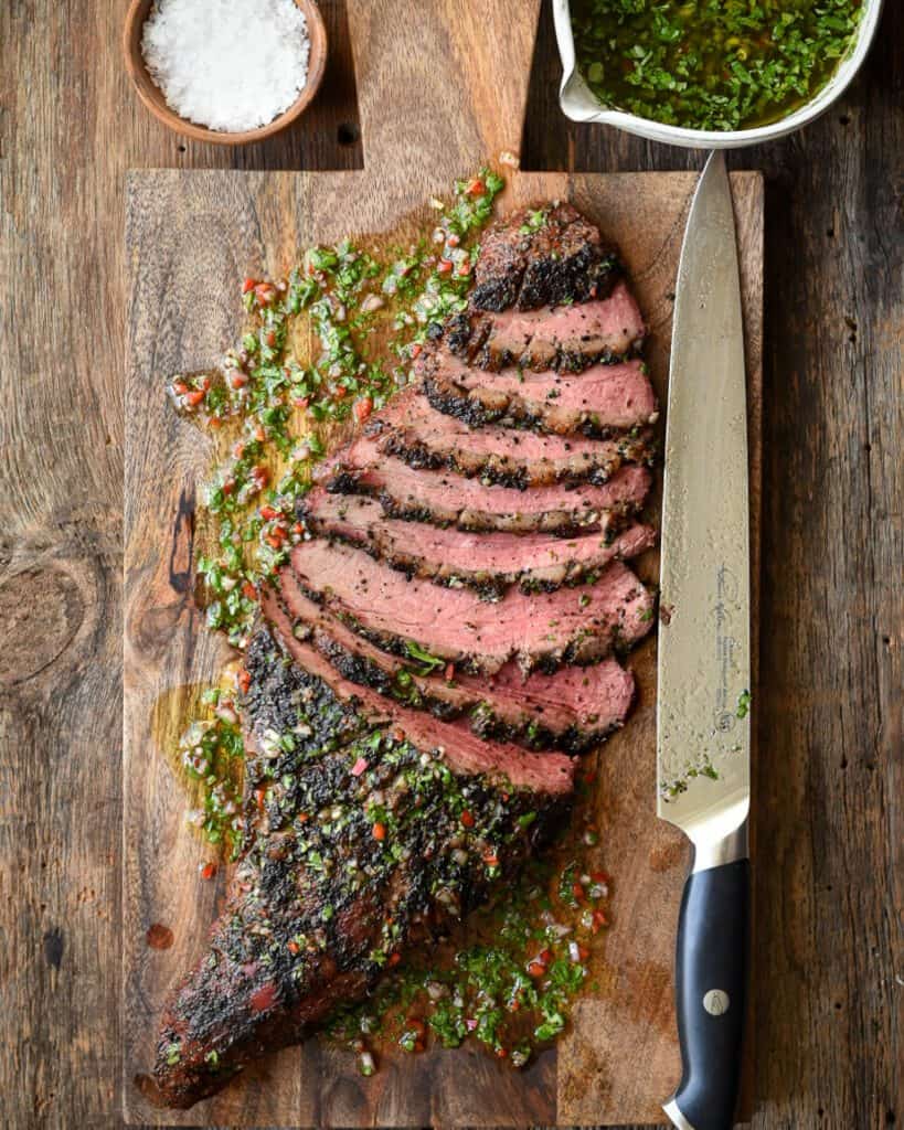A sliced Grilled Tri-Tip with Chimichurri on a board with a carving knife.