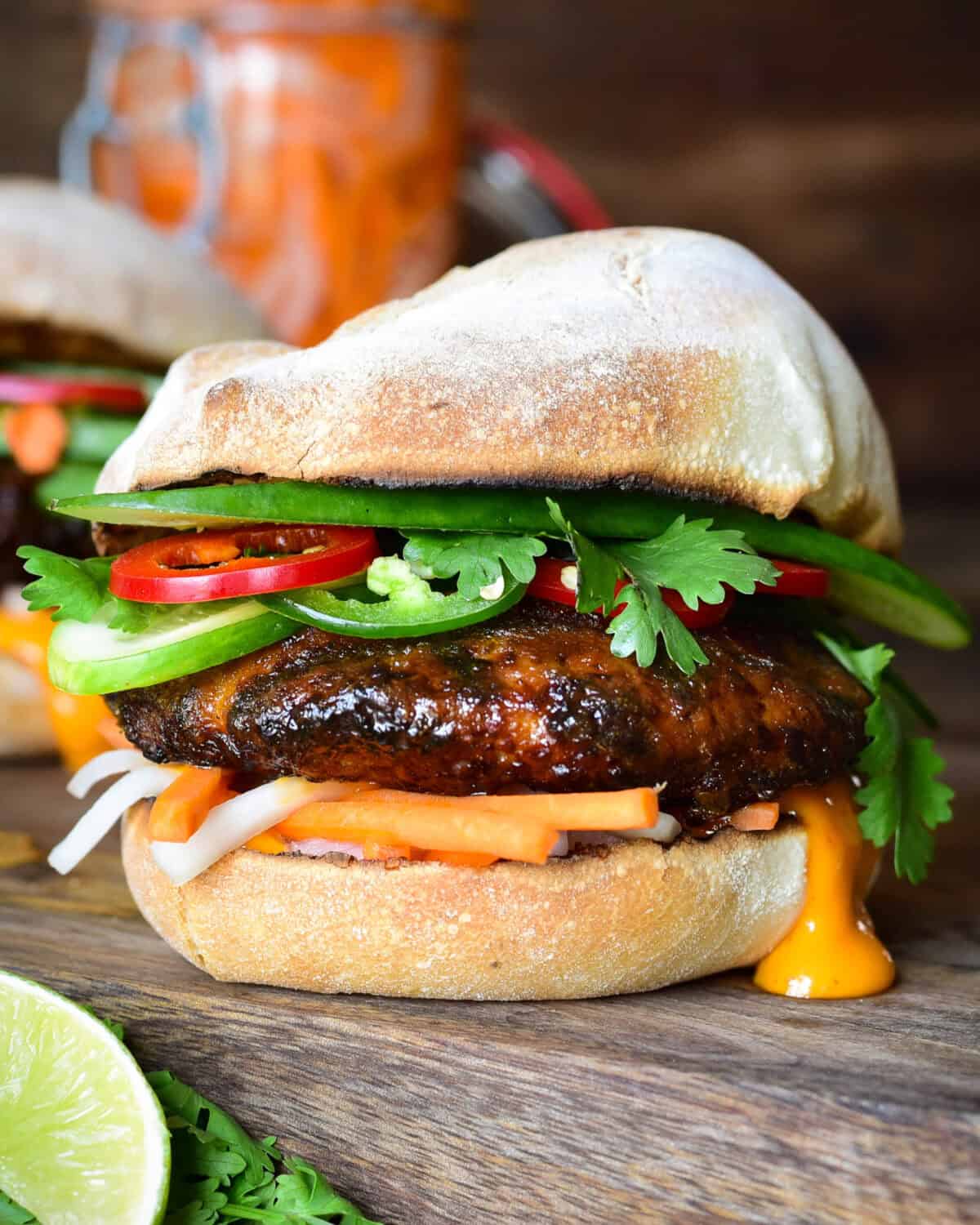 A side view of a Bánh Mì pork burger loaded with pickled and fresh vegetables and sauce on a French roll.