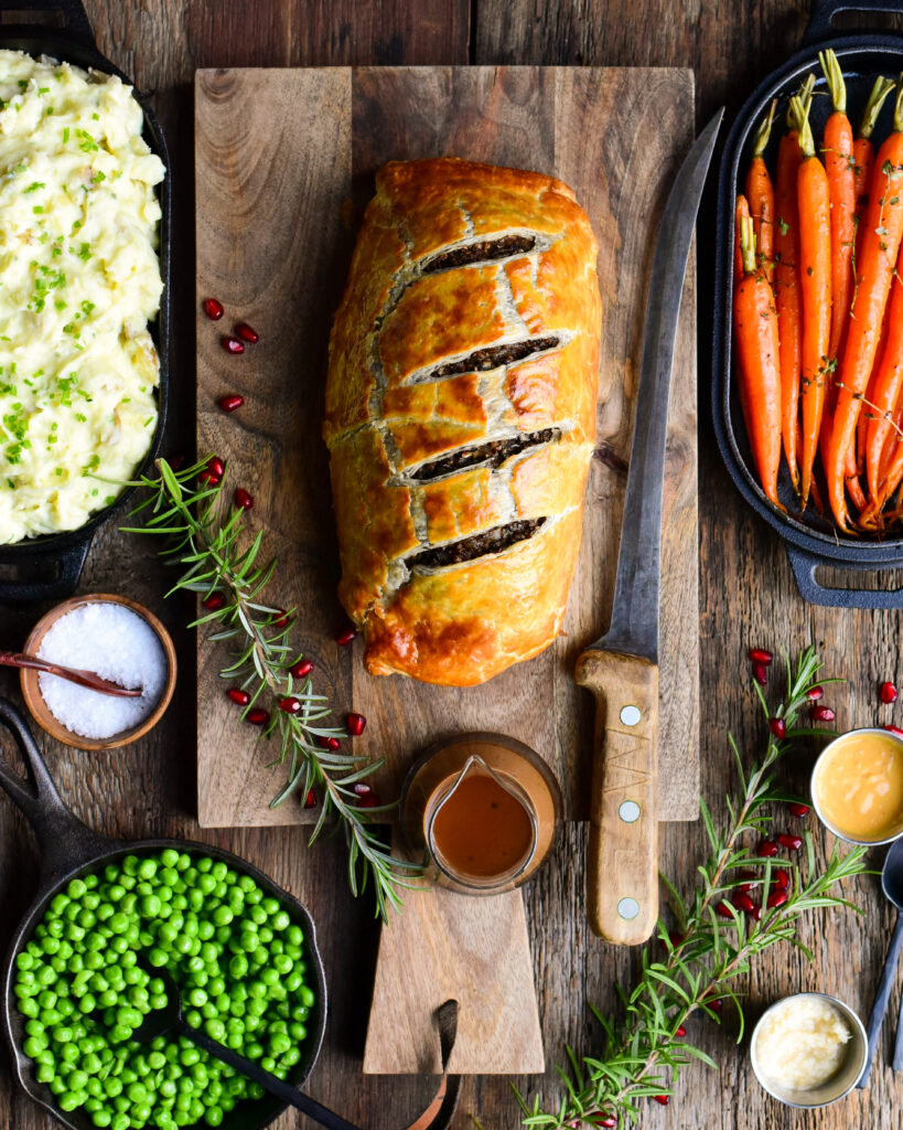 A top down image of a beef wellington on a wooden cutting board. This board is surrounded with suggested sides for beef wellington.