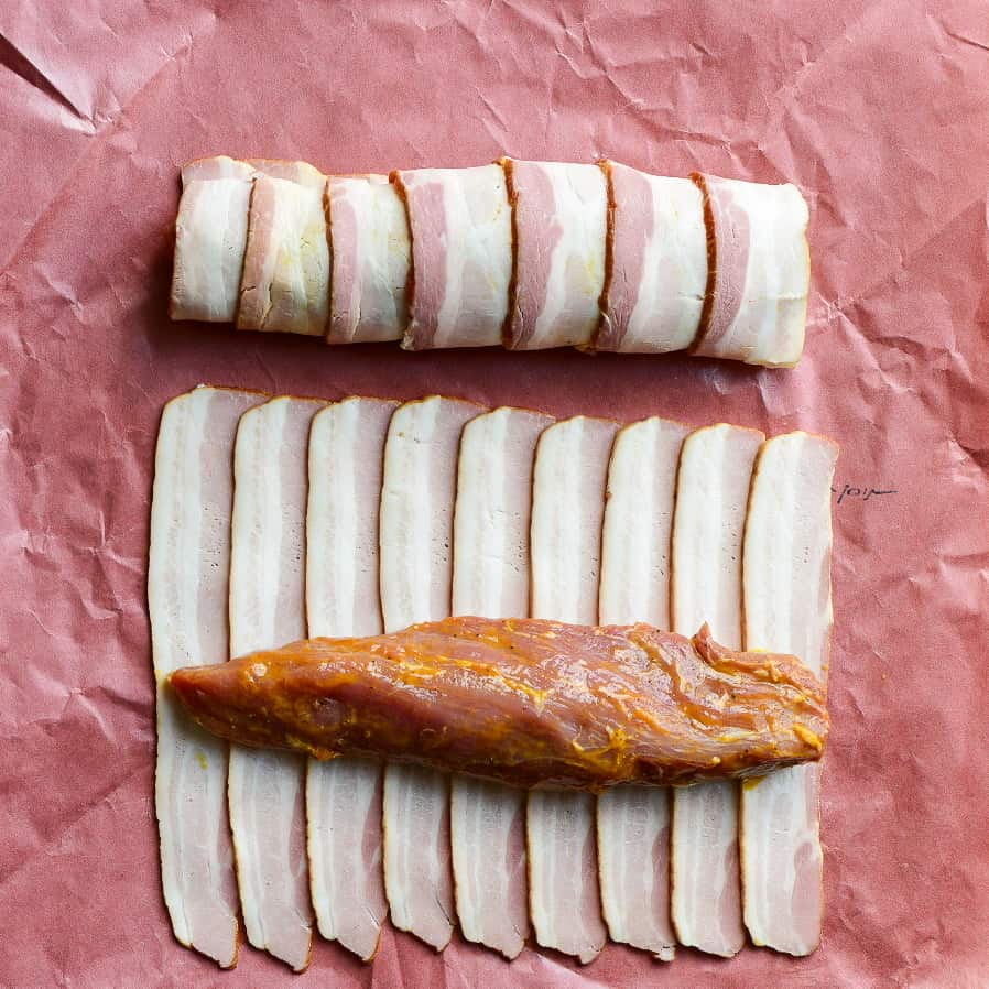 One pork tenderloin is wrapped neatly with bacon, and the other one is about to get wrapped in bacon.