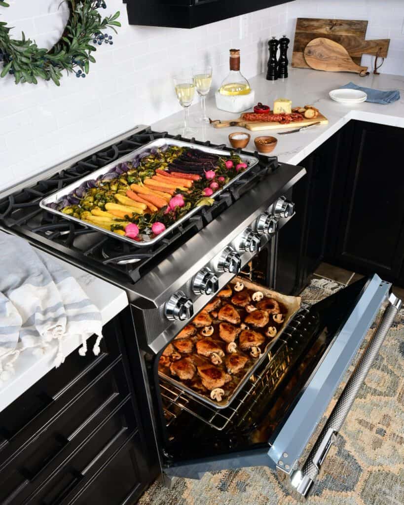 An image taken in a kitchen with the range open with a sheet pan of roasted chicken inside and a tray of colourful roasted vegetables on the top.