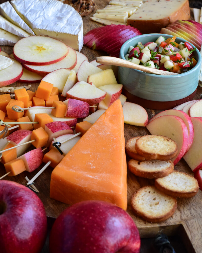 A close up od a cheese board with apples, a wedge of cheddar cheese, apple and cheese skewers, and a bowl of apple salsa.