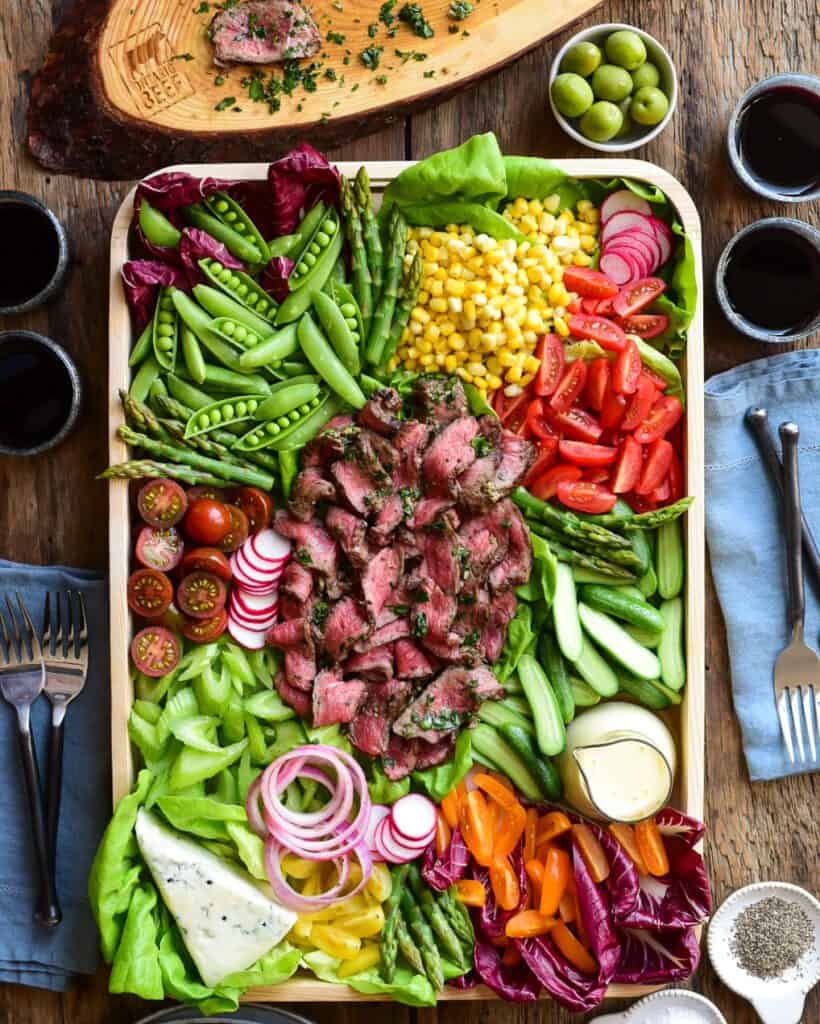 A bright and colourful grilled beef salad. Ribbons of medium rare beef are place in the middle of the tray and is surrounded by stack/piles of fresh vegetables, salad dressing and blue cheese.