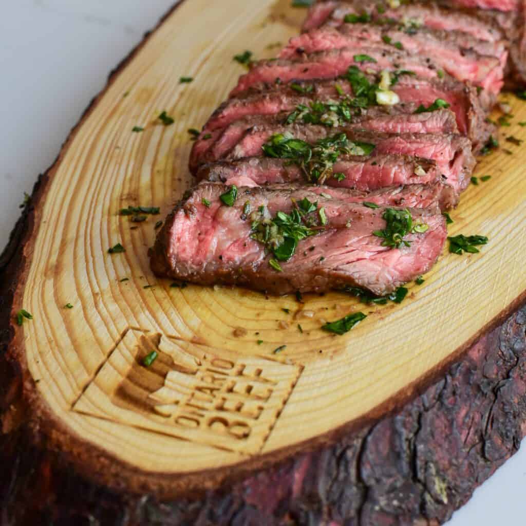 Medium rare slices of Ontario beef on a wood (bark still on the edges) board. The beef is resting in a board sauce and will be added to this grilled beef salad.
