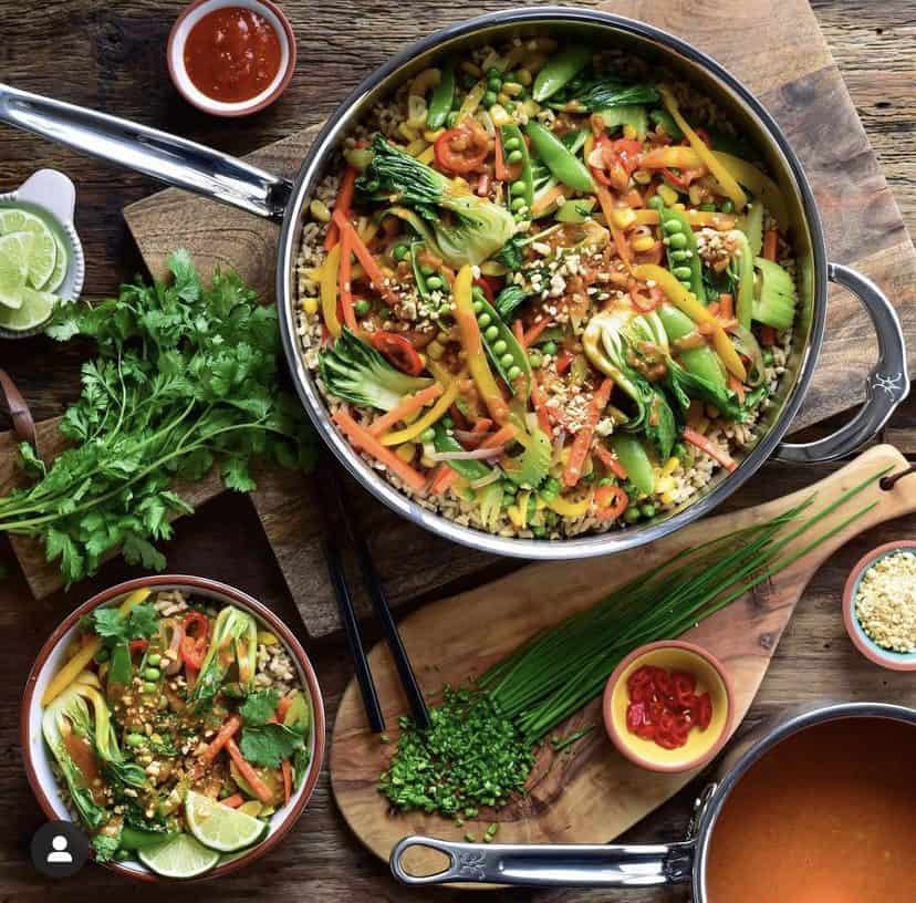 A top down image of a pan and a bowl of Red Thai curry peanut sauce and vegetables. Also layed out is cilantro, crushed peanuts, chopped chives and a pot of sauce.