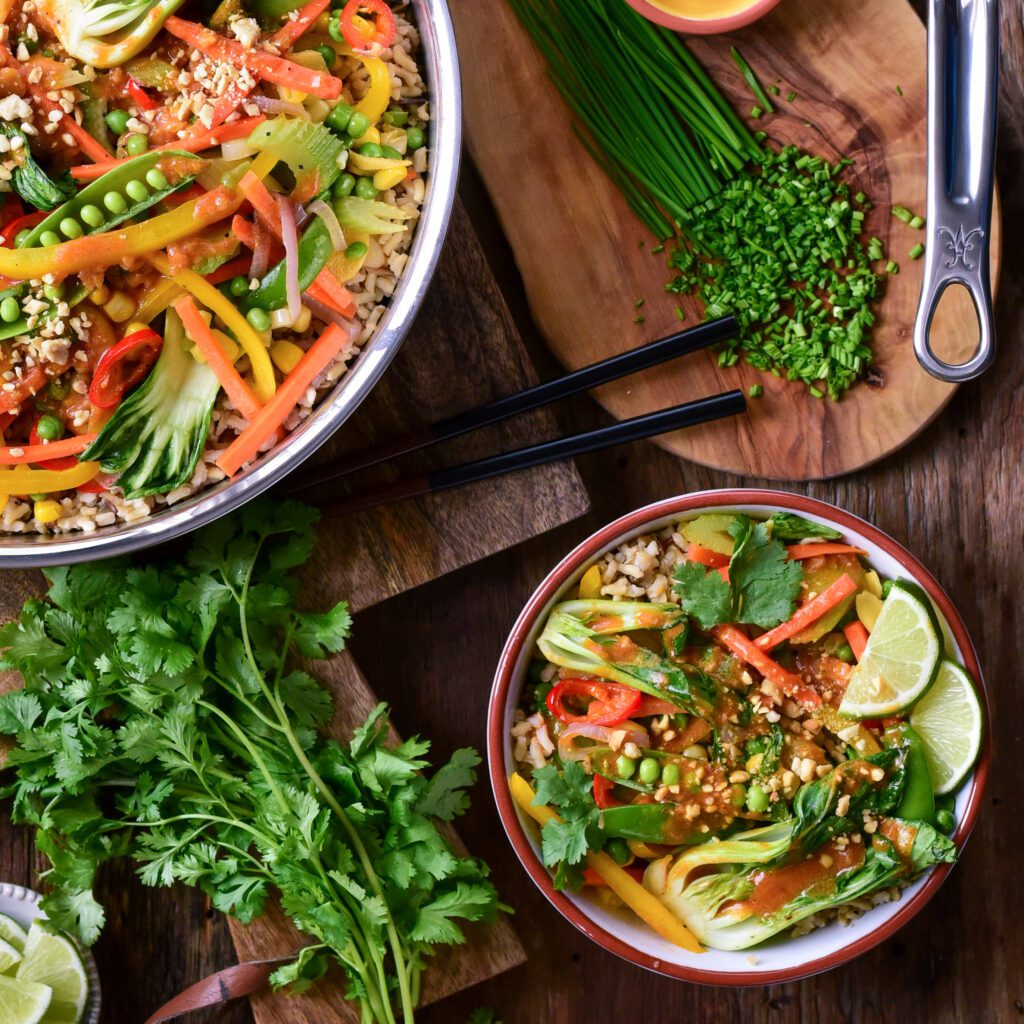 A top down image of a pan and a bowl of Red Thai curry peanut sauce and vegetables. Also layed out is cilantro, crushed peanuts, chopped chives and a pot of sauce.