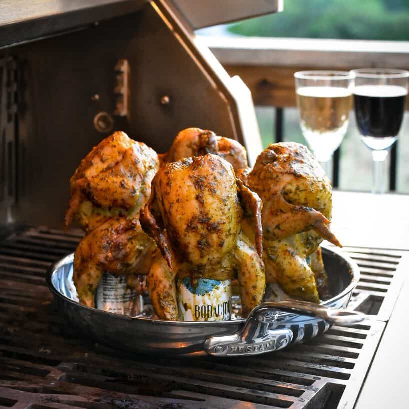 Four wine can Cornish hens are placed on a skillet on a grill. Two wine glasses (one white, one red) are in the background. 