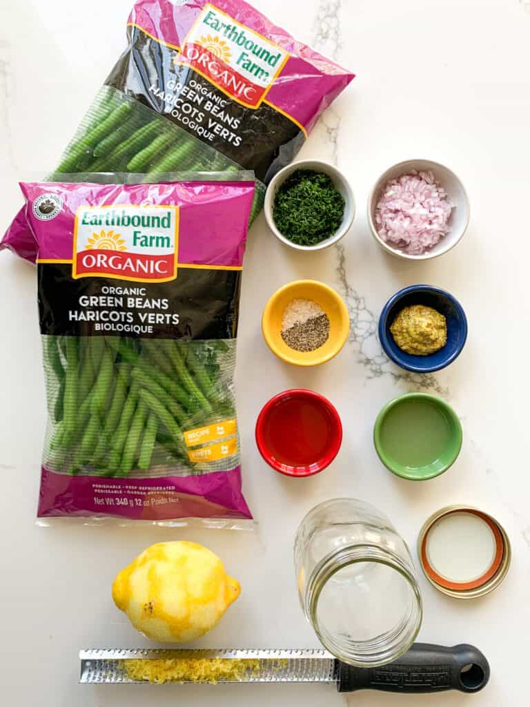 Ingredients for Earthbound Green Beans with Lemon, Dill, Dijon Dressing are portioned out in little colourful dishes beside two packages of beans.