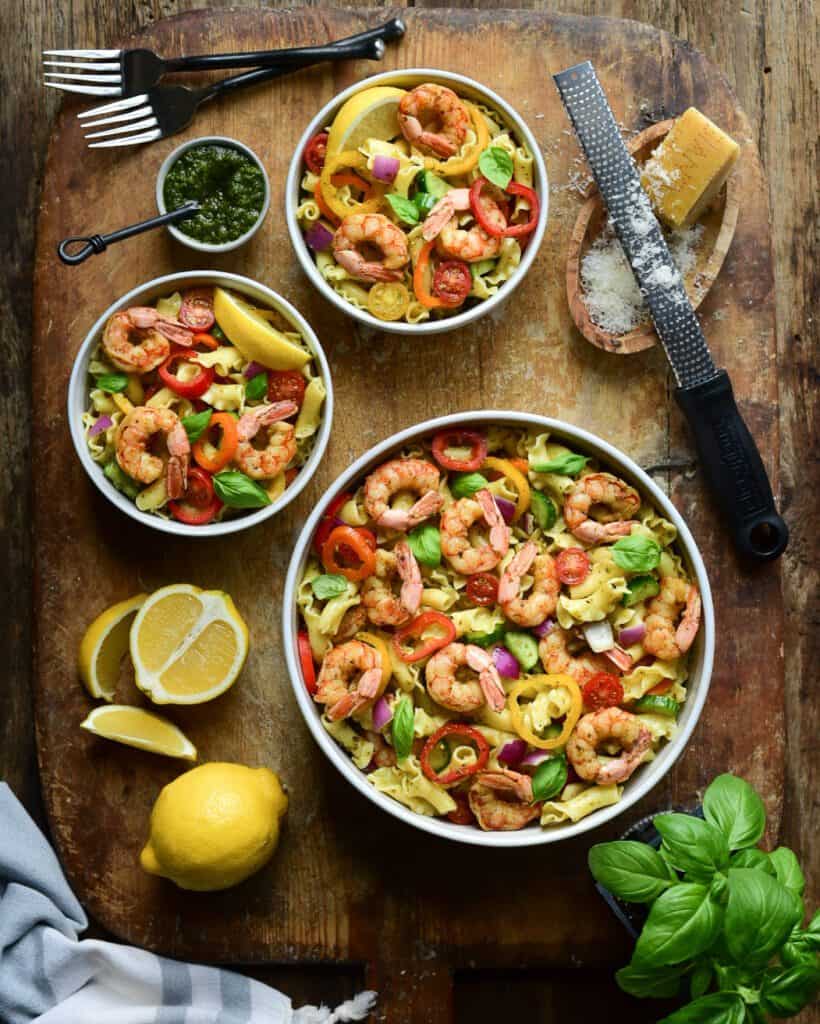 A large and two small bowls of shrimp pasta on a wooden cutting board. Also on this board are sliced lemons, basil, cheese and a grater.