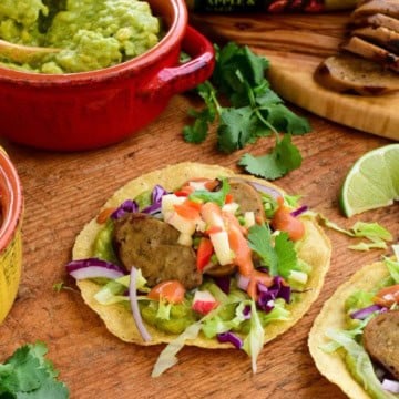 Close-up of apple and sage sausage tostadas topped with fresh vegetables and cilantro, served with guacamole and lime wedges on a wooden table.