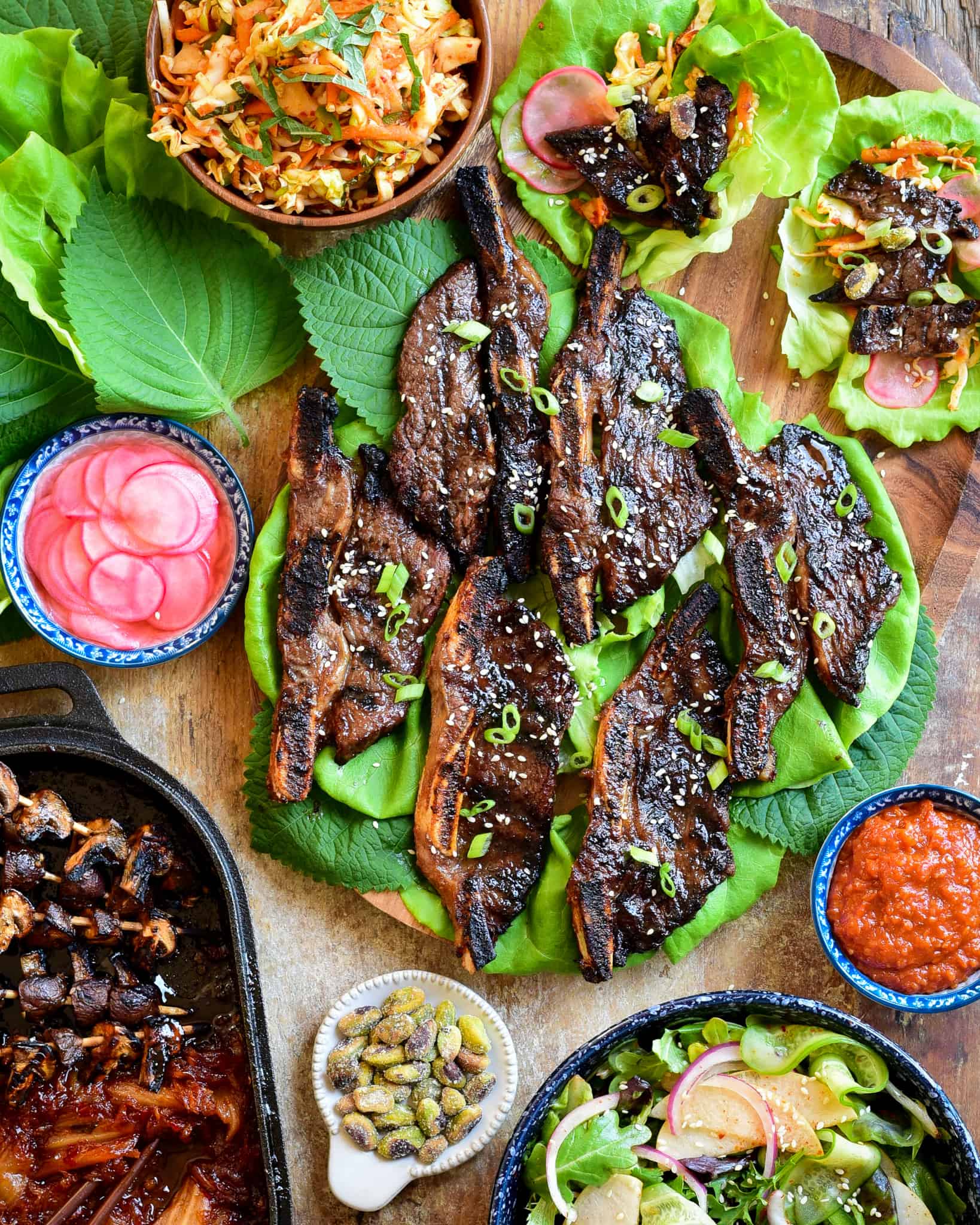 A Korean-Style Barbecue Beef Short Rib Lettuce Wraps spread on a board with coleslaw, salad, grilled mushrooms and kimchi.