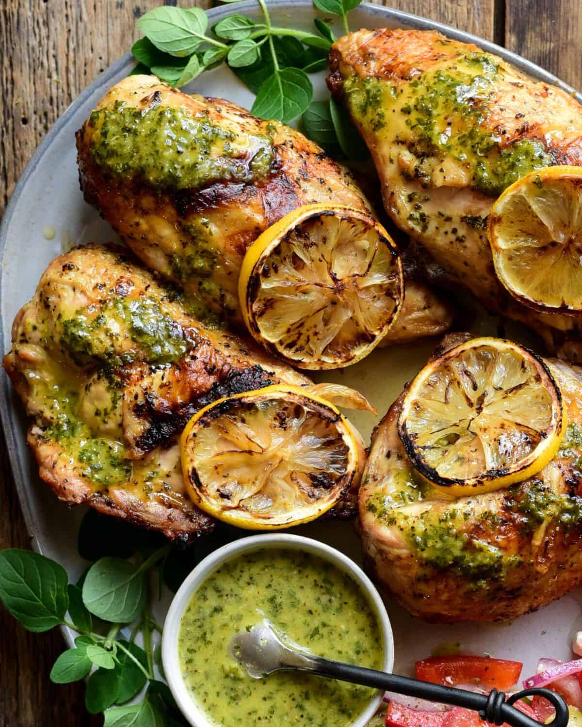 A close up of four grilled chicken breasts with a lemon-oregano sauce and grilled lemon slices and a bowl of sauce.