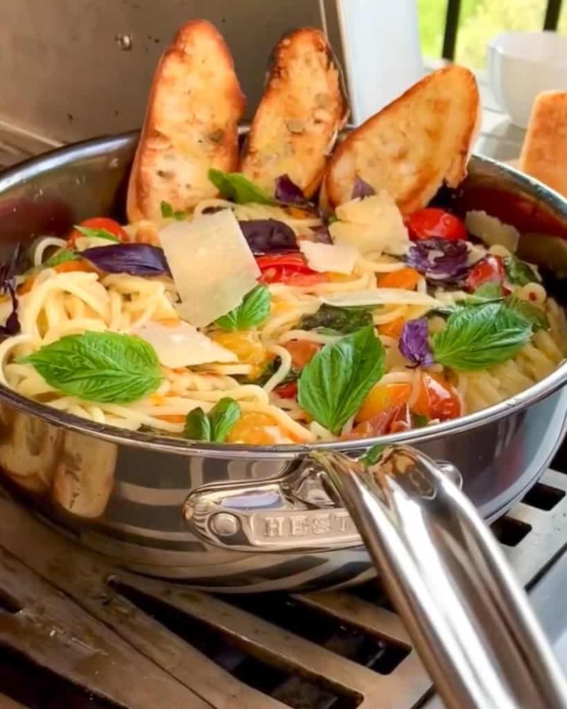 A large pan of cherry tomato spaghetti with garlic toast outside on a grill.