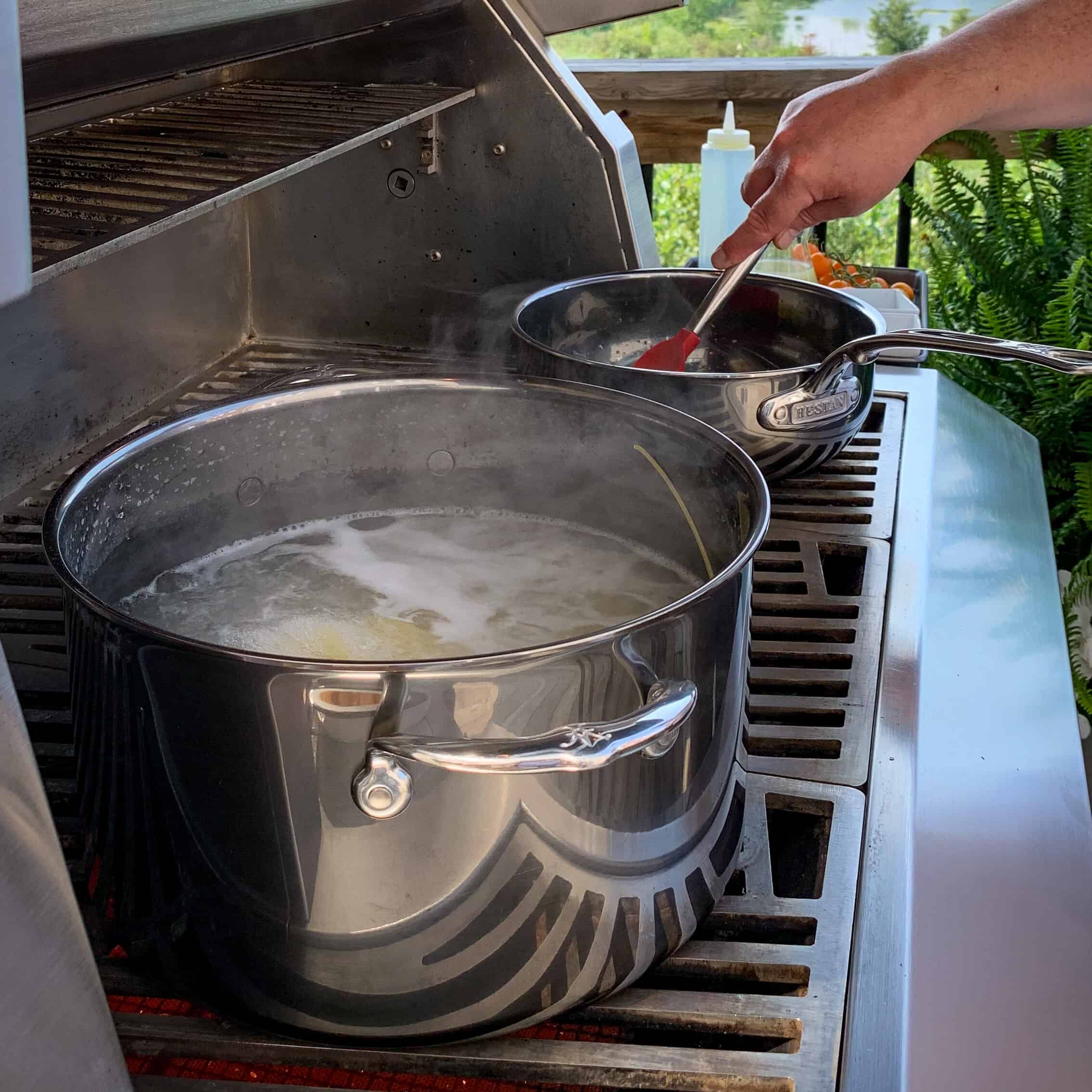 Two pots on the grill, one is boiling pasta the other is cooking the cherry tomato sauce.