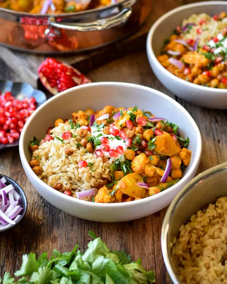 A bowl of curry topped with cilantro, arils and red onion. Topping, a copper pot and rice surround the bowl.