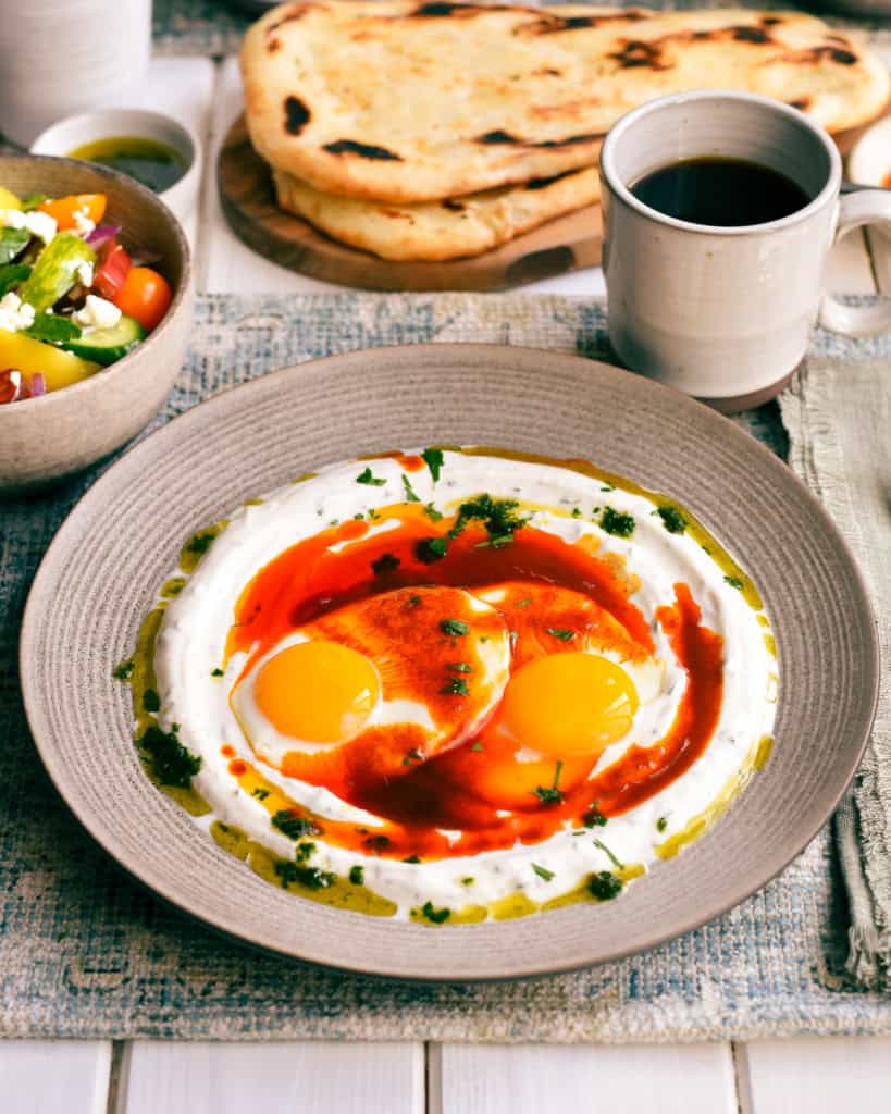 A plate of Turkish Turkish-inspired eggs (Çılbır) with pitas, salad and coffee on a table. 
