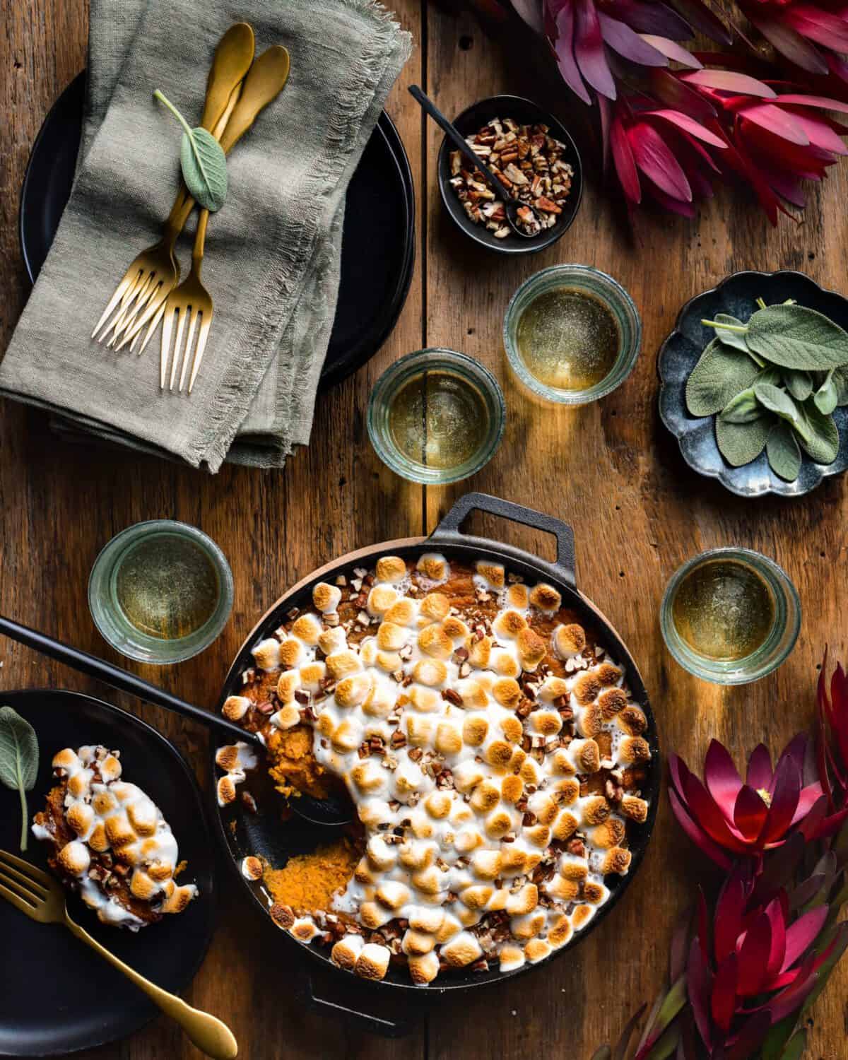 Sweet Potato Casserole with toasted marshmallows in a cast iron pan. A scoop is take out to reveal the inside and a portion is plated. Glassed, dishes, toppings and flowers surround the dish.
