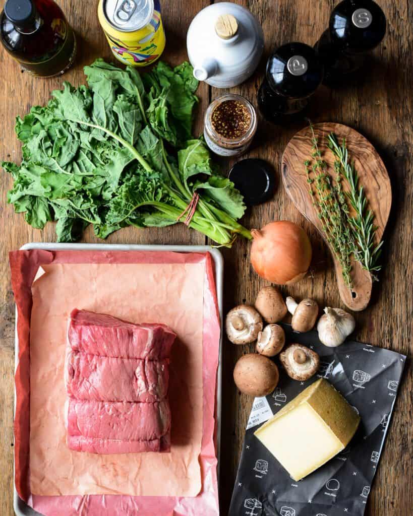 A top down image of a raw beef roast, cheese, mushrooms, onion, thyme, kale, mustard, olive oil, beer and salt and pepper.
