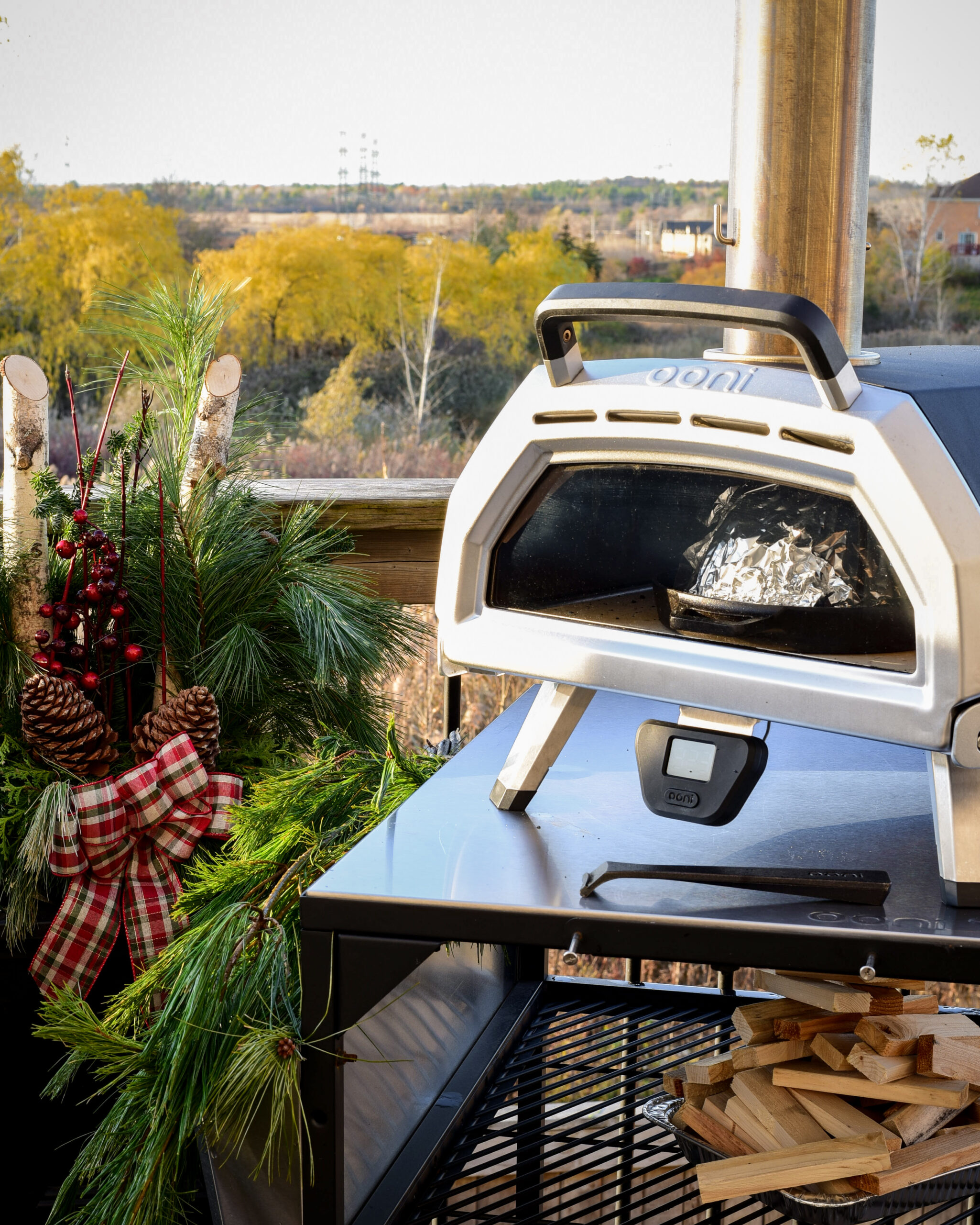 An Ooni oven on a table beside a festive planter with trees in the background.