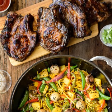 A top down image of a wok with a brightly coloured stir fry beside a cutting board with four beautifully charred pork chops.