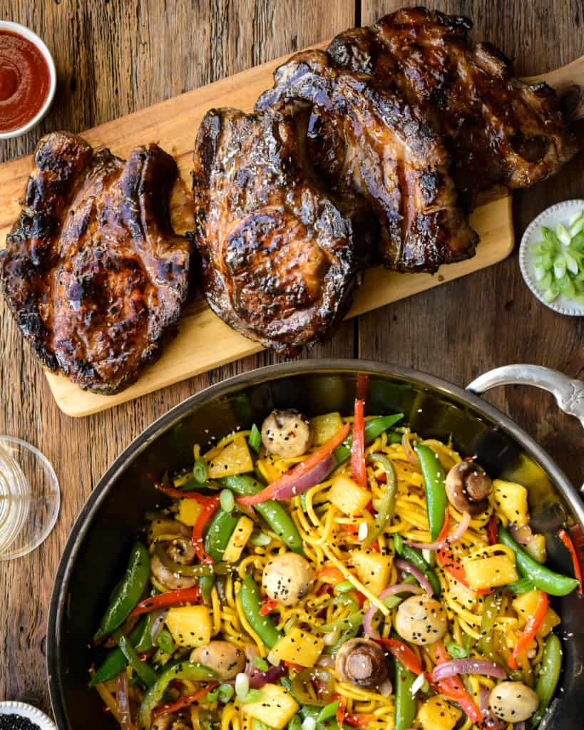 A top down image of a wok with a brightly coloured stir fry beside a cutting board with four beautifully charred pork chops.