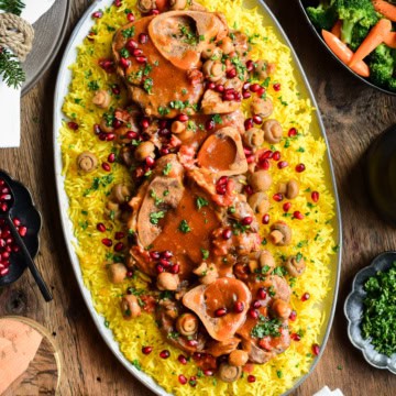 An overhead image of an oval platter with veal shanks and gravy over rice. Toppings and vegetables surround this platter.