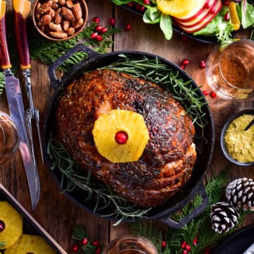 An image of a baked ham in a cast iron skillet with a pineapple on top. There is also winter salad, more baked pineapples, a carving knife set and holiday sprigs on the table.