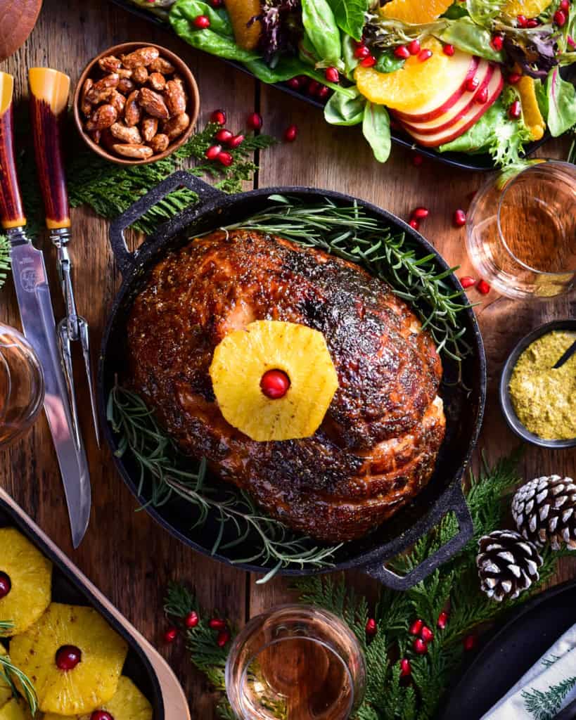 An image of a baked ham in a cast iron skillet with a pineapple on top. There is also winter salad, more baked pineapples, a carving knife set and holiday sprigs on the table.