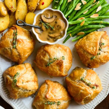 An oval platter with six individual ground beef wellingtons, a bowl of marsala mushroom sauce, potatoes and green beans.